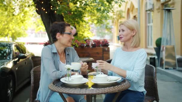 Ελκυστικές κυρίες φίλοι συνομιλούν σε υπαίθριο καφέ κρατώντας κούπες καφέ χαμογελαστά — Αρχείο Βίντεο