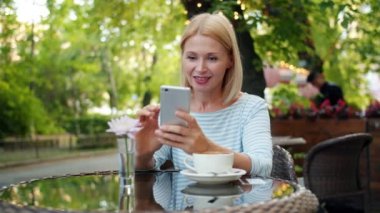 Kafe dışında akıllı telefon ekranı kullanan mutlu bir kadın yavaş çekim yapıyor.
