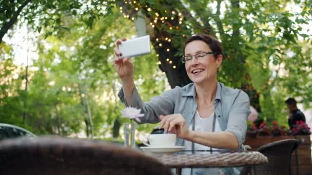 Ώριμη κυρία σε γυαλιά κάνοντας online κλήση βίντεο χαλαρωτικό στο Street Café στην πόλη — Αρχείο Βίντεο