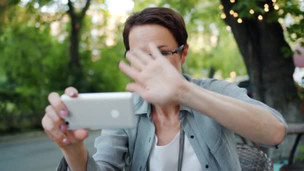 Mouvement lent de la femme faisant un appel vidéo en ligne à partir d'un café extérieur à l'aide d'un smartphone — Video