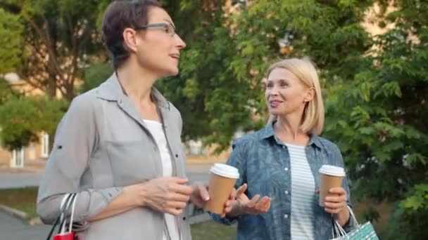 Alegre amigos caminando en parque charlando riendo sosteniendo café y bolsas — Vídeo de stock