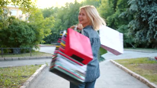 Cámara lenta de rubia madura girando afuera sosteniendo bolsas de compras divirtiéndose — Vídeos de Stock