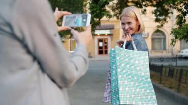 Mutlu shopaholic açık havada alışveriş çantaları tutan akıllı telefon kamera için poz