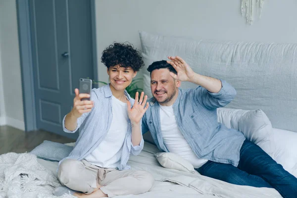 ベッドの上で手を振って話すスマートフォンでビデオ通話をする若いカップル — ストック写真