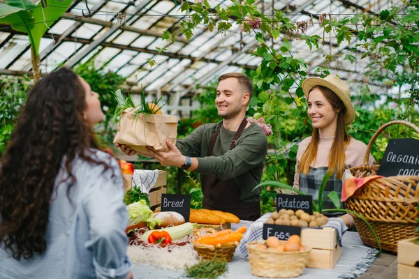 Pareja joven vendiendo alimentos orgánicos en el mercado agrícola en invernadero hablando con los clientes —  Fotos de Stock