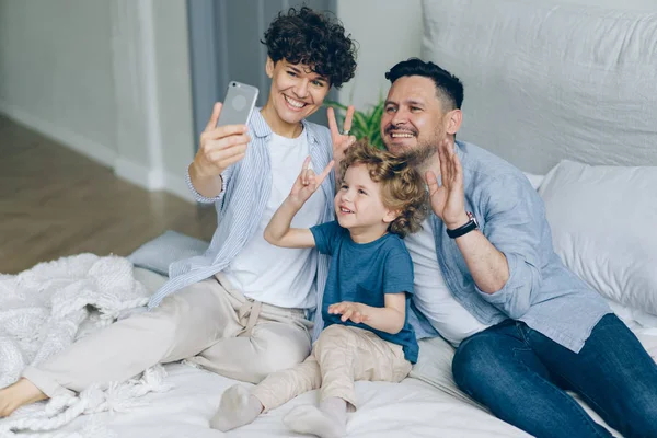 Madre padre hijo tomando selfie en la cama en casa posando para cámara de teléfono inteligente —  Fotos de Stock