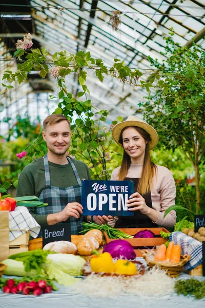 Двое счастливых фермеров держат открытую табличку, приветствуя клиентов на фермерском рынке — стоковое фото