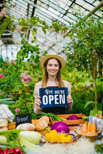 Bonita vendedora segurando sinal aberto no mercado agrícola sorrindo olhando para a câmera — Fotografia de Stock