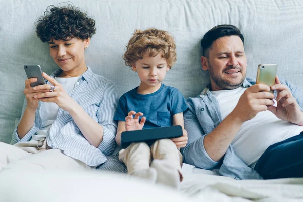 Homem e mulher usando smartphones enquanto criança jogando jogo no tablet na cama em casa — Fotografia de Stock