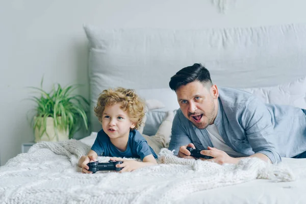 Padre e hijo jugando videojuego en la cama en casa presionando botones en el joystick —  Fotos de Stock