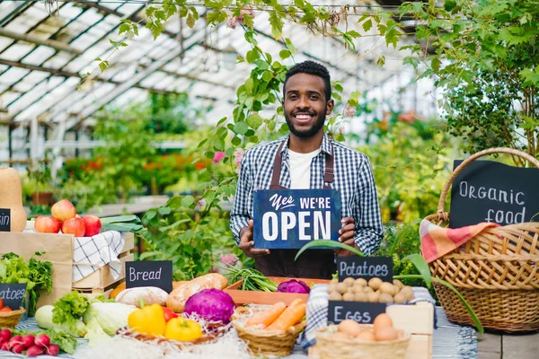 Продавец в фартуке держит открытый знак на рынке органических продуктов питания приветствуя людей — стоковое фото