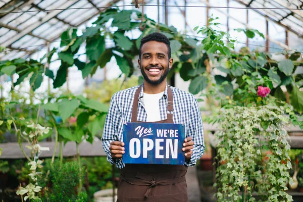 African American człowiek stoi w szklarni z jesteśmy otwarte znak uśmiechnięty — Zdjęcie stockowe