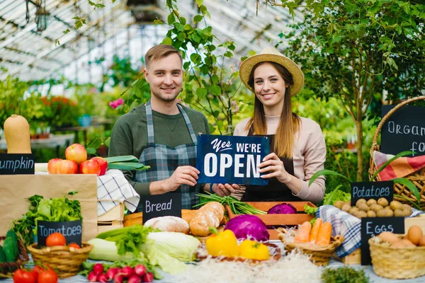 Парень и девушка держат открытую вывеску за столом с органическими продуктами на рынке — стоковое фото