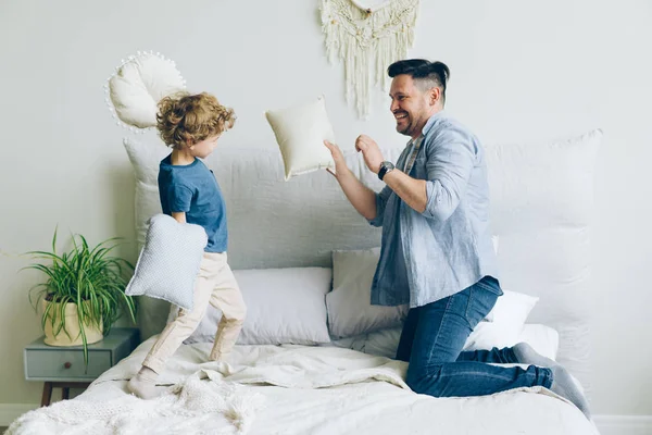 Joven hombre luchando almohadas con pequeño hijo divirtiéndose en la cama en el apartamento — Foto de Stock