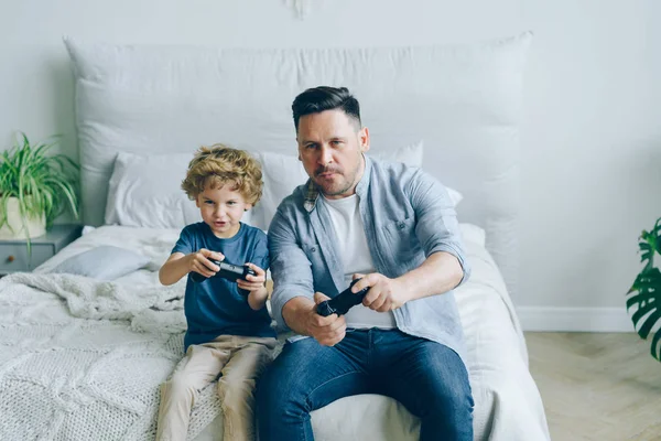 Lindo niño jugando videojuego con el padre sosteniendo joysticks divertirse en la cama — Foto de Stock