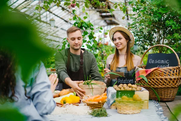 Продавец упаковывает овощи в бумажный пакет во время продажи на ферме, когда жена делает заметки — стоковое фото