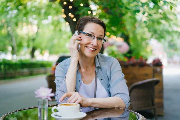 Зріла жінка в окулярах розмовляє на мобільному телефоні на відкритому повітрі кафе на відкритому повітрі — стокове фото
