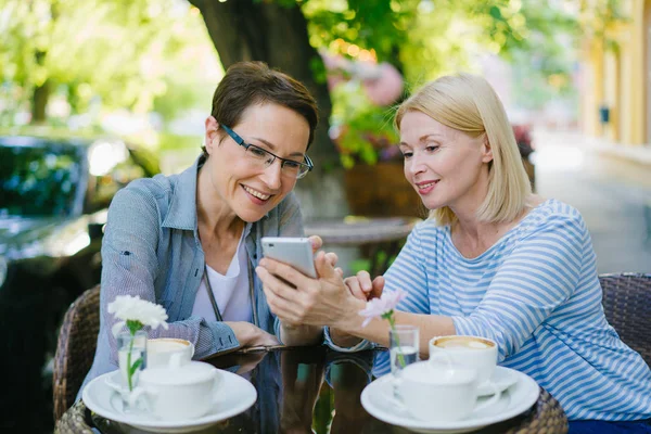 Hermosas mujeres maduras usando teléfono inteligente hablando riendo en la cafetería de la calle —  Fotos de Stock