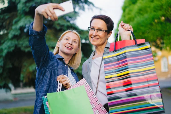 Vrouwen nemen selfie met smartphone camera poseren met boodschappentassen buitenshuis — Stockfoto