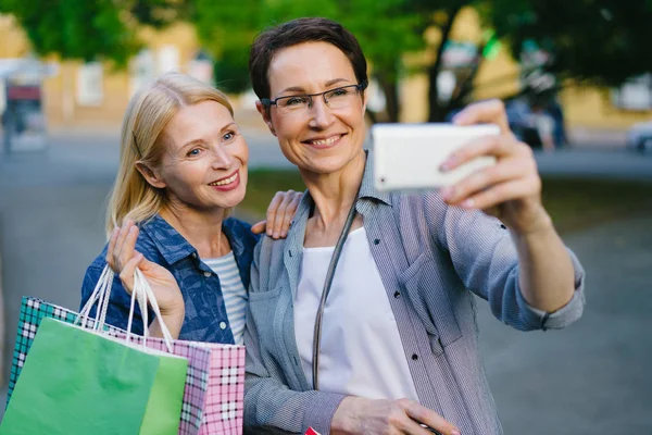 Vrouwelijke vrienden blonde en brunette nemen selfie met boodschappentassen buiten — Stockfoto