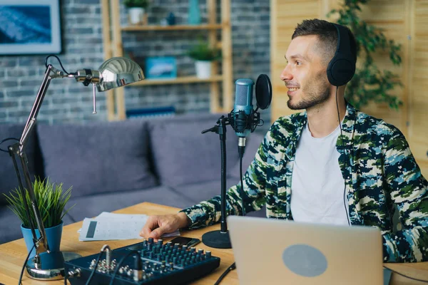 Fröhlicher Typ, der in Mikrofon spricht, Kopfhörer trägt und Laptop im Studio benutzt — Stockfoto