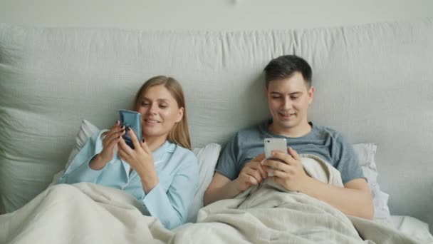 Jolie blonde utilisant un smartphone au lit partageant du contenu avec son mari souriant — Video