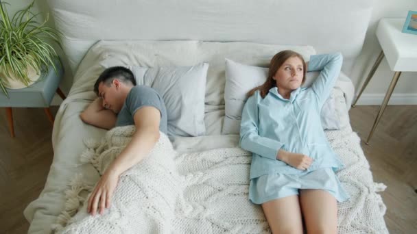 Sad girl in pajamas lying in bed sighing while guy boyfriend sleeping at home — Stock Video