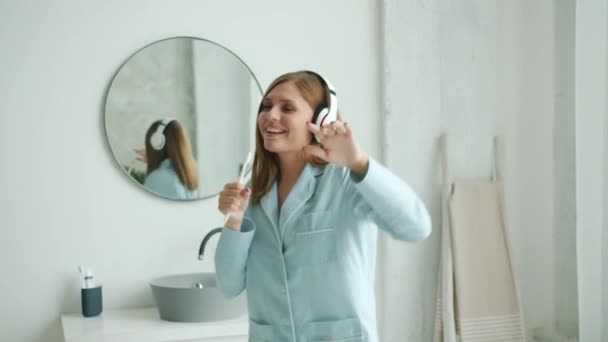 Chica alegre en los auriculares cepillarse los dientes cantando bailando en el baño en casa — Vídeo de stock