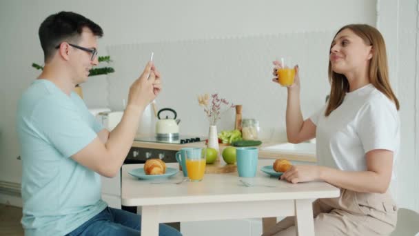 Mladá žena pózná ovocnou šťávou, zatímco manžel fotografi s smartphone — Stock video