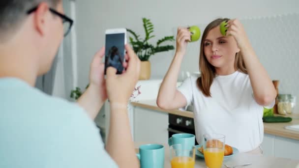 Żona pozowanie z jabłek podczas mąż biorąc zdjęcie ze smartfonem w kuchni — Wideo stockowe