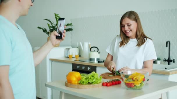 Dona de casa fazendo salada se divertindo enquanto o homem tira fotos com smartphone — Vídeo de Stock