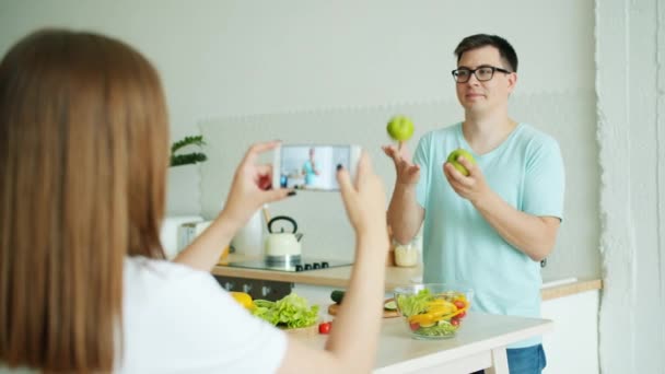 ガールフレンドがスマートフォンで写真を撮るとき、学生は台所でリンゴをジャグリング — ストック動画