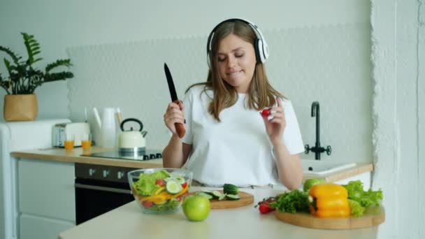 Mujer feliz en auriculares cocinando ensalada y bailando escuchando música en casa — Vídeos de Stock