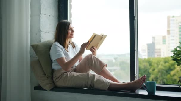 Beautiful girl reading book turning pages sitting on window-sill at home — Stock Video