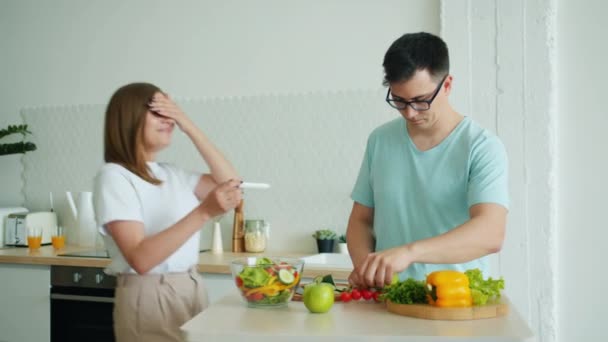 Мужчина муж готовит еду на кухне, когда счастливая девушка приносит тест на беременность — стоковое видео