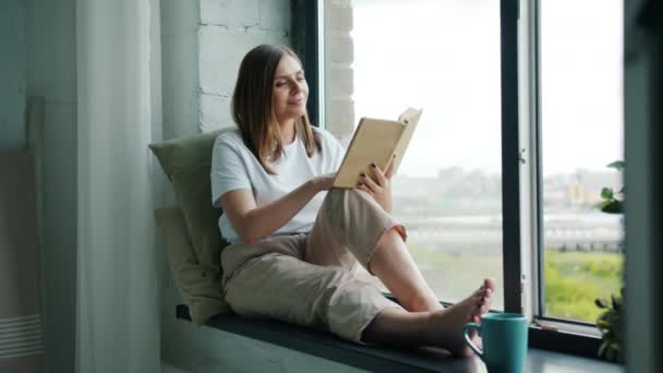 Bella giovane signora che legge libro sul davanzale della finestra e guardando fuori sorridente — Video Stock