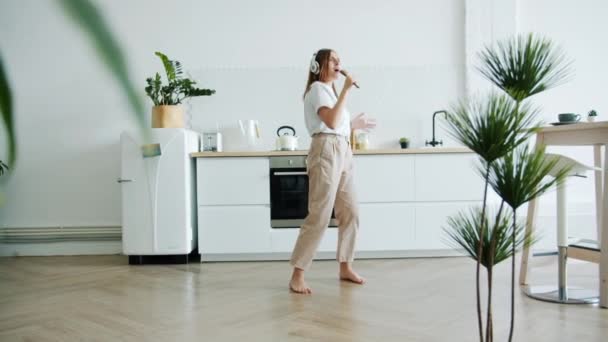 Woman in headphones dancing listening to music singing in spoon in kitchen — Stock Video