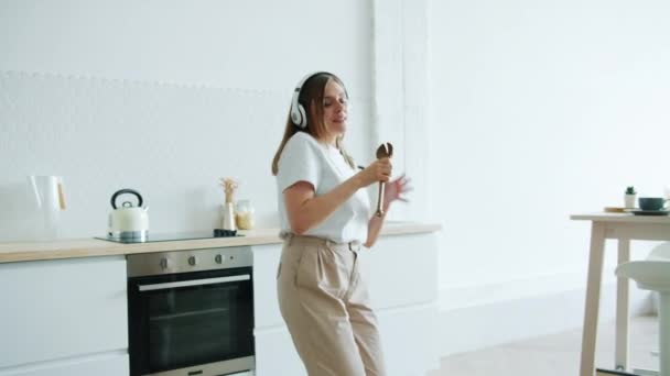 Alegre dama bailando cantando en cuchara escuchando música en auriculares en casa — Vídeo de stock