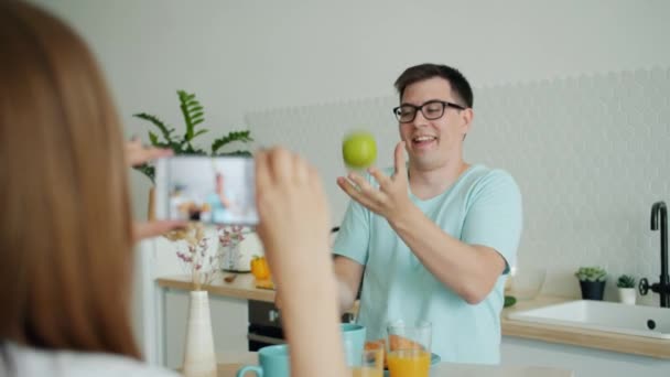 Hombre alegre haciendo malabarismos con frutas en la cocina mientras su esposa toma fotos con su teléfono inteligente — Vídeos de Stock