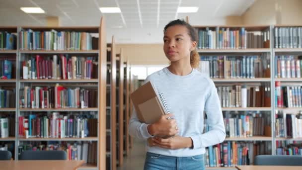 Menina afro-americana andando na biblioteca da universidade segurando livros olhando ao redor — Vídeo de Stock