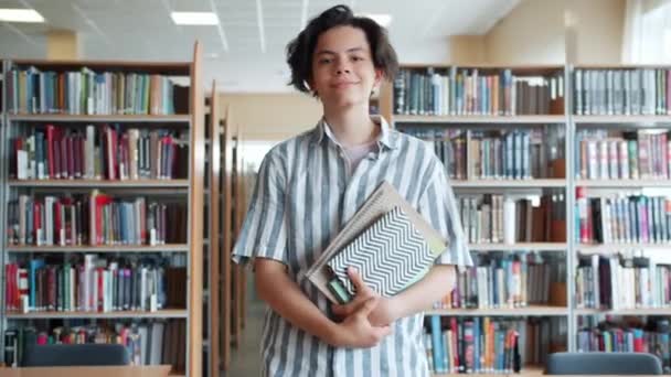 Glad tonåring går i College bibliotek hålla böcker titta på kamera — Stockvideo