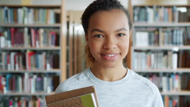 Porträt einer attraktiven afrikanisch-amerikanischen Studentin, die lächelnd Bücher in der Bibliothek hält — Stockvideo