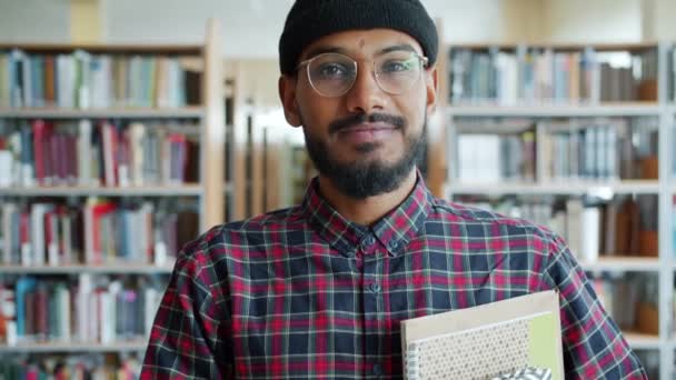 Retrato de un afroamericano barbudo con sombrero sosteniendo libros en la biblioteca sonriendo — Vídeos de Stock