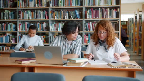 Estudiantes que estudian en la biblioteca hablando usando portátil haciendo investigación juntos — Vídeos de Stock