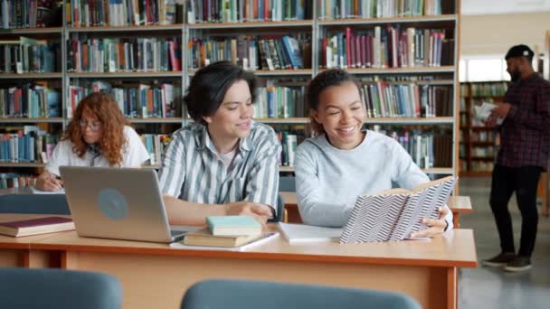 Chica alegre y chico estudiando en la biblioteca de la universidad hablando con libros para portátiles — Vídeos de Stock