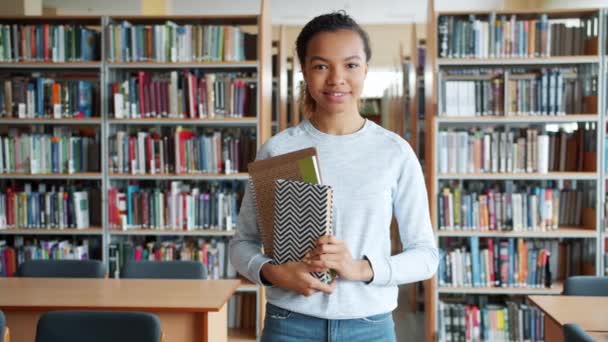 Афроамериканка, стоящая в библиотеке с книгами, улыбающимися в камеру — стоковое видео