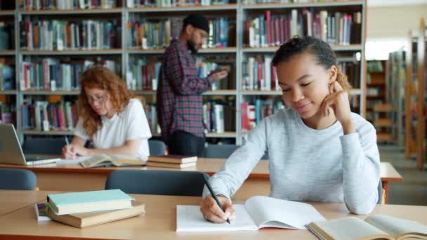 Menina afro-americana escrevendo na biblioteca, enquanto as pessoas andando no fundo — Vídeo de Stock