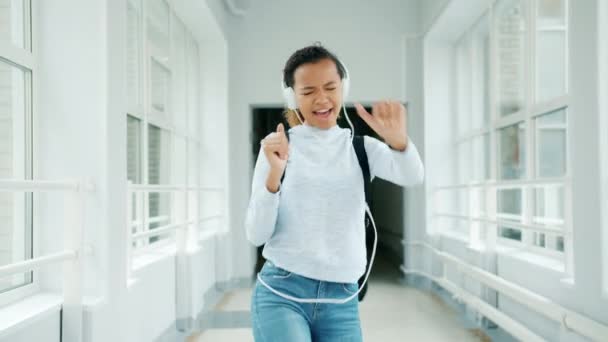 Chica alegre en auriculares divirtiéndose en el salón de la universidad cantando baile — Vídeos de Stock