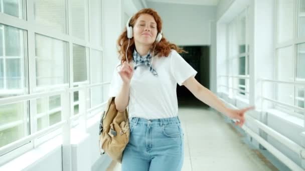 Гарненька руда в навушниках танцює в шкільному вестибюлі, розважаючись з музикою — стокове відео