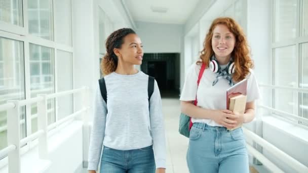 Chicas amigas caminando en la sala de la escuela sosteniendo libros charlando haciendo chistes — Vídeo de stock